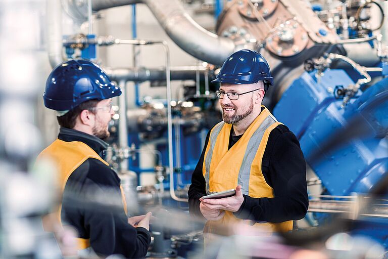 Dois técnicos industriais, com capacete azul e colete refletor, verificam dados num tablet digital em frente às máquinas de uma unidade de produção. A avaliação correta da conformidade exige determinar se o equipamento requer uma inspeção por parte de um organismo notificado ou um controlo interno da produção.
